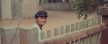 Movie still from “Burning Patience” (2022), directed by Rodrigo Sepúlveda – A young man wearing a hat standing in front of a fence; Close Up shot, Over the shoulder angle