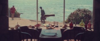 Movie still from “Burning Patience” (2022), directed by Rodrigo Sepúlveda – A man standing at a table near the water; Wide shot, Over the shoulder angle