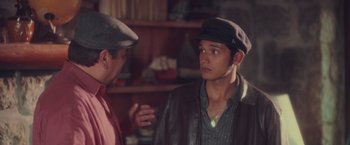 Movie still from “Burning Patience” (2022), directed by Rodrigo Sepúlveda – A man wearing a hat talking to another man in front of a shelf; Close Up shot, Over the shoulder angle