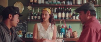 Movie still from “Burning Patience” (2022), directed by Rodrigo Sepúlveda – A woman sitting in front of a shelf in a room; Medium shot, Over the shoulder angle
