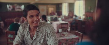 Movie still from “Burning Patience” (2022), directed by Rodrigo Sepúlveda – A woman smiles while sitting at a table in a restaurant; Close Up shot, Over the shoulder angle