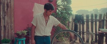 Movie still from “Burning Patience” (2022), directed by Rodrigo Sepúlveda – A man in a white shirt and blue jeans holding a bicycle tire; Medium shot, Low angle