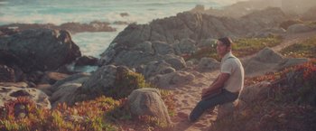 Movie still from “Burning Patience” (2022), directed by Rodrigo Sepúlveda – A man sitting on the beach next to the ocean; Wide shot, High angle