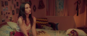 Movie still from “Burning Patience” (2022), directed by Rodrigo Sepúlveda – A woman sitting on top of a bed next to papers; Medium shot, Over the shoulder angle