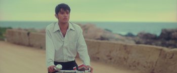 Movie still from “Burning Patience” (2022), directed by Rodrigo Sepúlveda – A young man riding a bike on top of a dirt road; Medium shot, Low angle