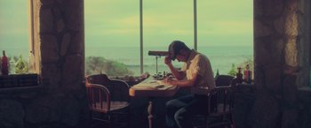 Movie still from “Burning Patience” (2022), directed by Rodrigo Sepúlveda – A man sitting at a table looking at a book; Medium shot, High angle