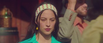 Movie still from “Burning Patience” (2022), directed by Rodrigo Sepúlveda – A young woman with a polka dot headband and earrings; Close Up shot, Over the shoulder angle