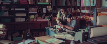 Movie still from “Burning Patience” (2022), directed by Rodrigo Sepúlveda – A man and a boy working in a room full of books; Medium shot, High angle