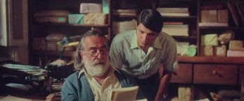 Movie still from “Burning Patience” (2022), directed by Rodrigo Sepúlveda – A man reading a book to another man in a room; Medium shot, Over the shoulder angle