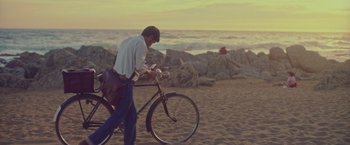 Movie still from “Burning Patience” (2022), directed by Rodrigo Sepúlveda – A man on a bicycle on the beach at sunset; Wide shot, Low angle