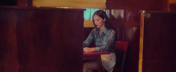 Movie still from “Burning Patience” (2022), directed by Rodrigo Sepúlveda – A woman sitting at a table in front of a laptop computer; Medium shot, Low angle