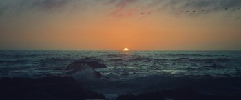 Movie still from “Burning Patience” (2022), directed by Rodrigo Sepúlveda – The sun is setting over the ocean with a flock of birds flying above it; Extreme Wide shot, High angle