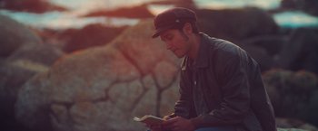 Movie still from “Burning Patience” (2022), directed by Rodrigo Sepúlveda – A man sitting on the ground while reading a book; Medium shot, High angle
