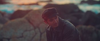 Movie still from “Burning Patience” (2022), directed by Rodrigo Sepúlveda – A man wearing a hat sitting on the ground in front of some rocks; Close Up shot, High angle