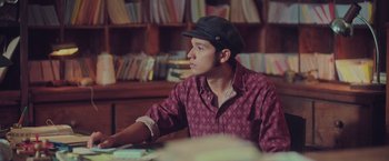 Movie still from “Burning Patience” (2022), directed by Rodrigo Sepúlveda – A young man wearing a hat sitting at a table; Medium shot, Low angle