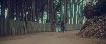 Movie still from “Burning Patience” (2022), directed by Rodrigo Sepúlveda – A man riding a bike on a dirt road near trees; Extreme Wide shot, Low angle
