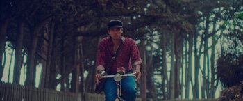 Movie still from “Burning Patience” (2022), directed by Rodrigo Sepúlveda – A man riding a bike in a park with trees in the background; Medium shot, Low angle