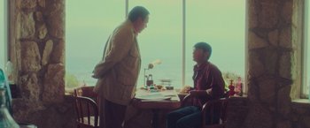 Movie still from “Burning Patience” (2022), directed by Rodrigo Sepúlveda – Two men sitting at a table with food in front of a window; Medium shot, Over the shoulder angle