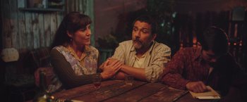 Movie still from “Burning Patience” (2022), directed by Rodrigo Sepúlveda – A man and a woman sitting at a wooden table; Medium shot, High angle