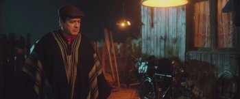Movie still from “Burning Patience” (2022), directed by Rodrigo Sepúlveda – A man wearing a blanket and a hat standing in front of a wooden fence; Medium shot, Low angle