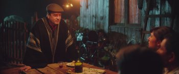 Movie still from “Burning Patience” (2022), directed by Rodrigo Sepúlveda – A man sitting at a wooden table with a bottle of wine; Medium shot, Over the shoulder angle