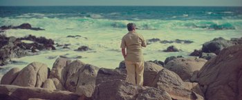 Movie still from “Burning Patience” (2022), directed by Rodrigo Sepúlveda – An older man standing on top of rocks looking out at the ocean; Wide shot, High angle