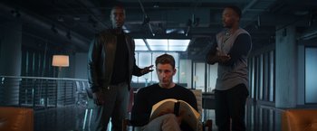 Movie still from “Captain America: Civil War” (2016), directed by Anthony Russo – A man sitting in a chair reading a book while two other men look on; Medium shot, Low angle