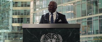 Movie still from “Captain America: Civil War” (2016), directed by Anthony Russo – A man in a suit and tie at a podium in front of a building; Medium shot, Low angle