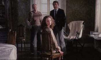 Movie still from “The Sacrifice” (1986), directed by Andrei Tarkovsky – A girl sitting in a chair between two men in a room; Wide shot, High angle