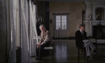 Movie still from “The Sacrifice” (1986), directed by Andrei Tarkovsky – A man sitting on a chair in a living room next to another man; Wide shot, High angle