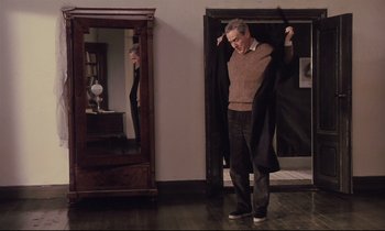 Movie still from “The Sacrifice” (1986), directed by Andrei Tarkovsky – A man standing in front of a mirror holding a jacket; Wide shot, Over the shoulder angle