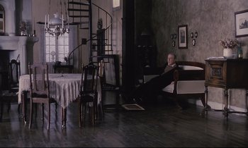 Movie still from “The Sacrifice” (1986), directed by Andrei Tarkovsky – An older man sitting on a couch in a living room; Wide shot, High angle