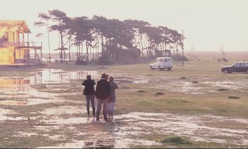 Movie still from “The Sacrifice” (1986), directed by Andrei Tarkovsky – Three people are standing in a field with a van in the background; Wide shot, High angle