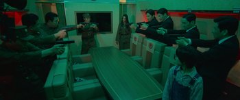 Movie still from “Carter” (2022), directed by Jung Byung-gil – A group of people standing around a table in a room; Medium shot, High angle