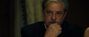Movie still from “Casino Royale” (2006), directed by Martin Campbell – An older man in a black suit and white shirt is holding his hand to his face; Close Up shot, Over the shoulder angle