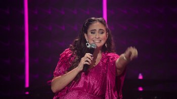 Movie still from “Catherine Cohen: The Twist...? She's Gorgeous” (2022), directed by Steven Brill – A woman in a pink dress is holding a microphone; Medium shot, High angle