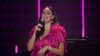 Movie still from “Catherine Cohen: The Twist...? She's Gorgeous” (2022), directed by Steven Brill – A woman in a pink dress singing into a microphone; Medium shot, Low angle