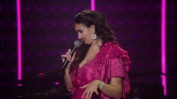 Movie still from “Catherine Cohen: The Twist...? She's Gorgeous” (2022), directed by Steven Brill – A woman in a pink dress is holding a microphone; Medium shot, High angle