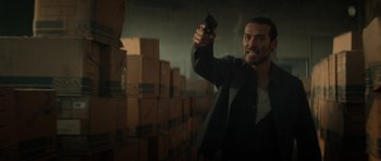 Movie still from “Centaur” (2022), directed by Daniel Calparsoro – A man holding up a cell phone in a room filled with boxes; Medium shot, Low angle
