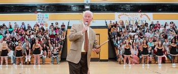 Movie still from “Central Intelligence” (2016), directed by Rawson Marshall Thurber – An older man is holding a microphone in front of a crowd; Medium shot, Low angle