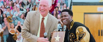 Movie still from “Central Intelligence” (2016), directed by Rawson Marshall Thurber – A man holding a microphone next to a man holding a plaque; Medium shot, Over the shoulder angle