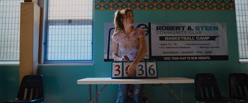 Movie still from “Champions” (2023), directed by Bobby Farrelly – A woman standing next to a table holding a sign; Medium shot, Low angle