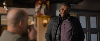 Movie still from “Champions” (2023), directed by Bobby Farrelly – An older man wearing a black jacket standing in front of a painting; Medium shot, Over the shoulder angle