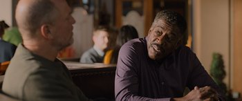 Movie still from “Champions” (2023), directed by Bobby Farrelly – An older man sitting at a table in front of other people; Close Up shot, Over the shoulder angle