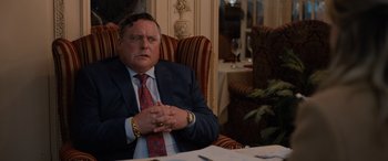 Movie still from “Champions” (2023), directed by Bobby Farrelly – A man in a suit and tie sitting in a chair in front of a table; Close Up shot, Over the shoulder angle