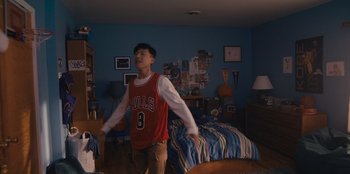 Movie still from “Chang Can Dunk” (2023), directed by Jingyi Shao – A young man in a room with a bed and blue walls; Wide shot, Over the shoulder angle