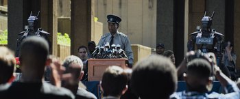 Movie still from “Chappie” (2015), directed by Neill Blomkamp – A man in a uniform speaking at a podium; Medium shot, Over the shoulder angle