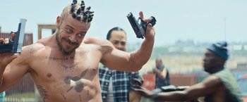 Movie still from “Chappie” (2015), directed by Neill Blomkamp – A shirtless man holding up a gun in front of a group of people; Close Up shot, Over the shoulder angle