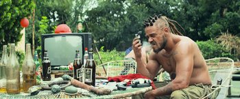 Movie still from “Chappie” (2015), directed by Neill Blomkamp – A man with tattoos and hair is sitting at a table with beer; Medium shot, Low angle