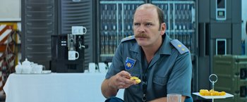 Movie still from “Chappie” (2015), directed by Neill Blomkamp – A police officer eating a donut while sitting down; Close Up shot, Over the shoulder angle